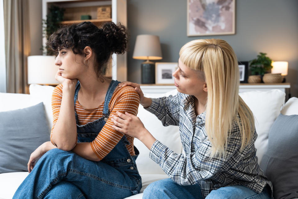 Two women seated on a couch: one appears upset, looking away, while the other, with blond hair, comforts her by placing a hand on her shoulder