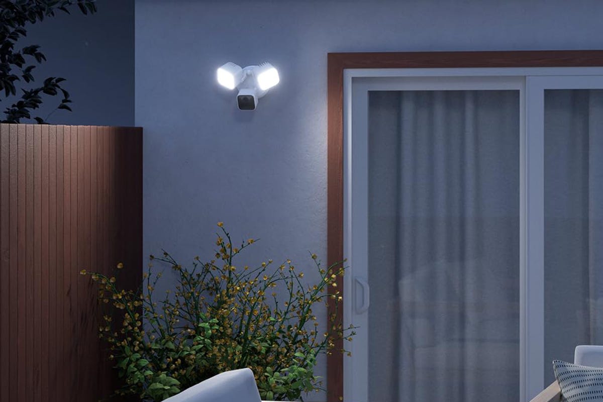 A Blink floodlight cam perched on a stucco wall beside a glass door at night.