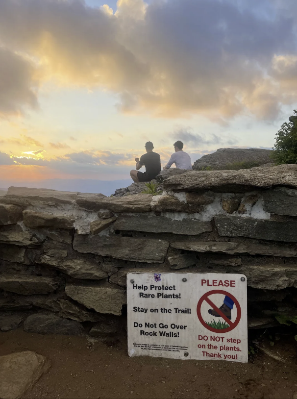 Two people sit on a rock wall overlooking a sunset. A sign reads: "Help Protect Rare Plants! Stay on the Trail! Do Not Go Over Rock Walls! Please DO NOT step on the plants. Thank you!"