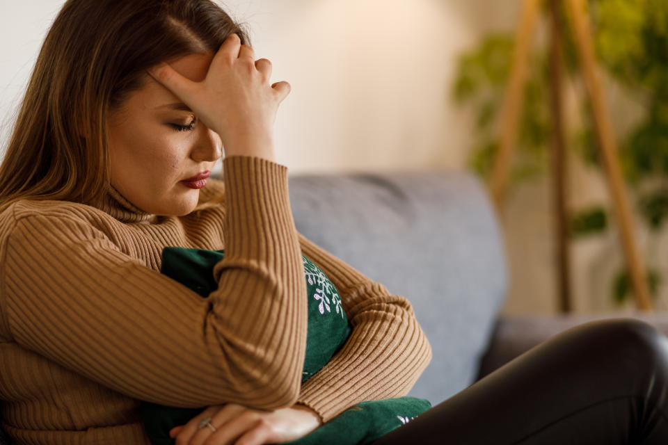 A woman sits on a couch, leaned forward with her hand on her forehead, appearing stressed or deep in thought