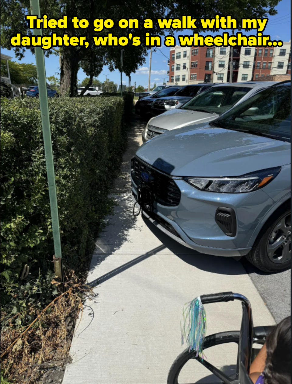 A blue car parked partially on the sidewalk, blocking the path. A person holding the handle of a wheelchair is visible in the lower right. Trees and buildings in the background
