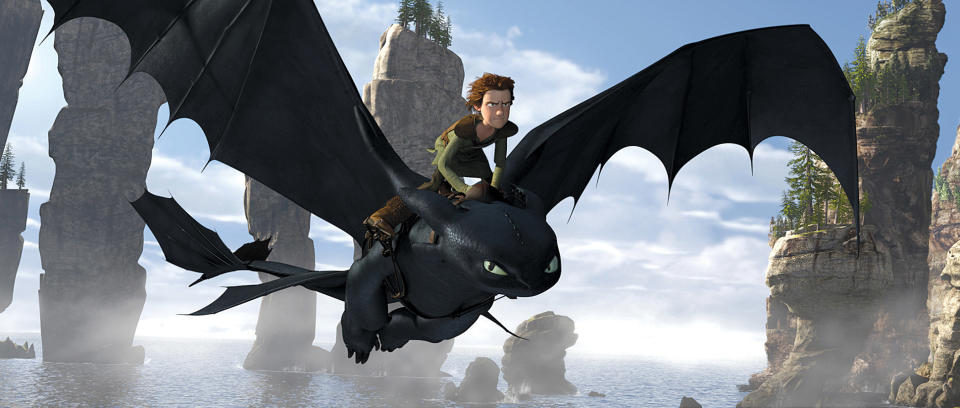 Hiccup rides Toothless, a black dragon, over a rocky ocean landscape in a scene from "How to Train Your Dragon."