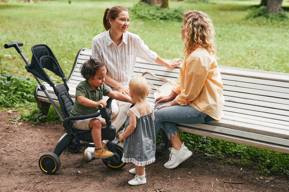 Two women sitting on a park bench chatting, one with a toddler in a stroller, the other with a standing toddler