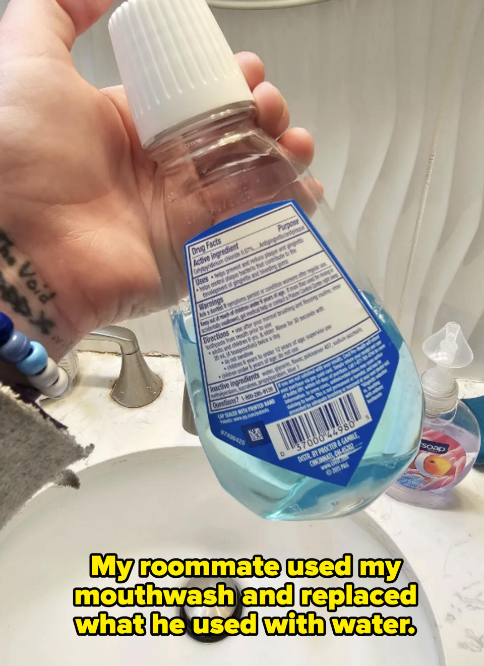A hand holding a bottle of mouthwash above a sink. The bottle displays drug facts, active ingredients, directions, and a barcode