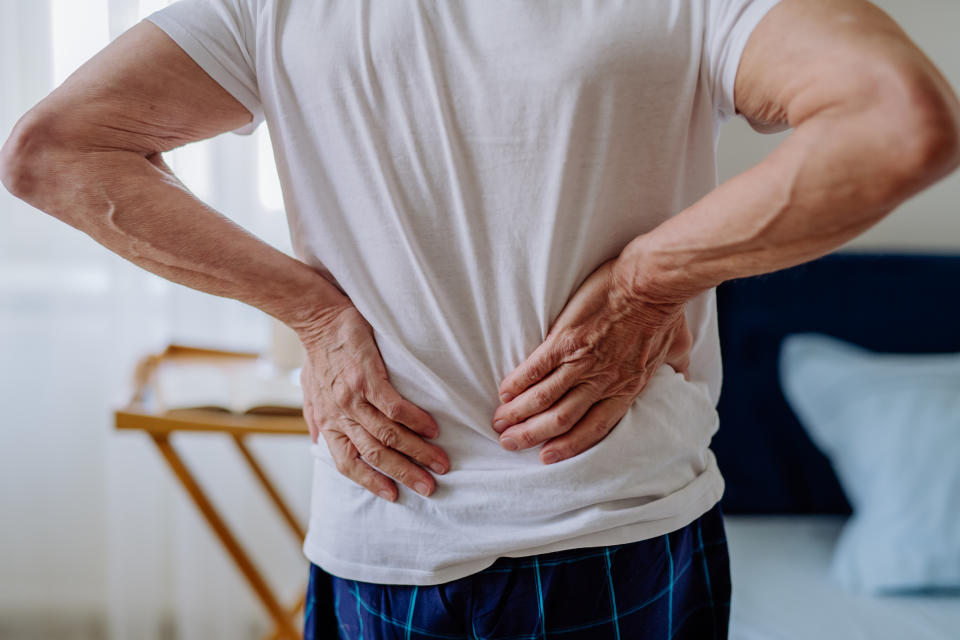 A person in a white t-shirt and checkered pants is holding their lower back, indicating back pain. The background shows a bedroom scene with a bed and nightstand