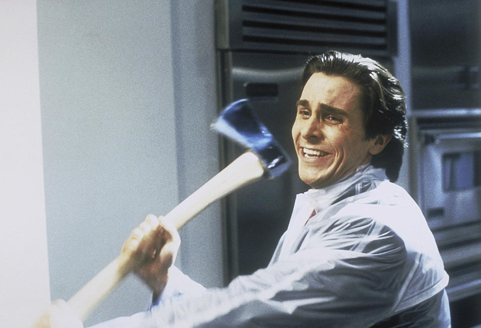 Christian Bale, wearing a clear raincoat over a white shirt, holds an axe with a menacing expression in a kitchen scene from "American Psycho."