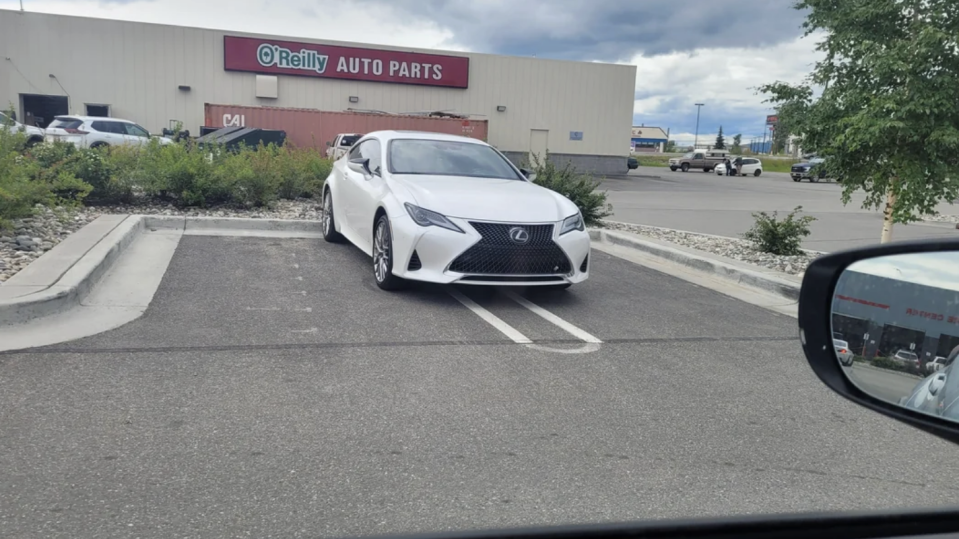 A white Lexus is parked over two spaces in a parking lot outside an O'Reilly Auto Parts store. The side mirror of another car is visible in the foreground