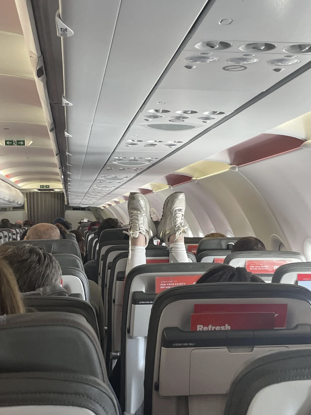 Passengers seated in an airplane with one person's feet wearing sneakers raised in the air, positioned on the headrests of the seats in front of them