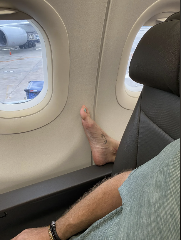 An airplane passenger's foot rests against the window, with their leg extending under the armrest. The view outside shows part of the airport tarmac and another aircraft