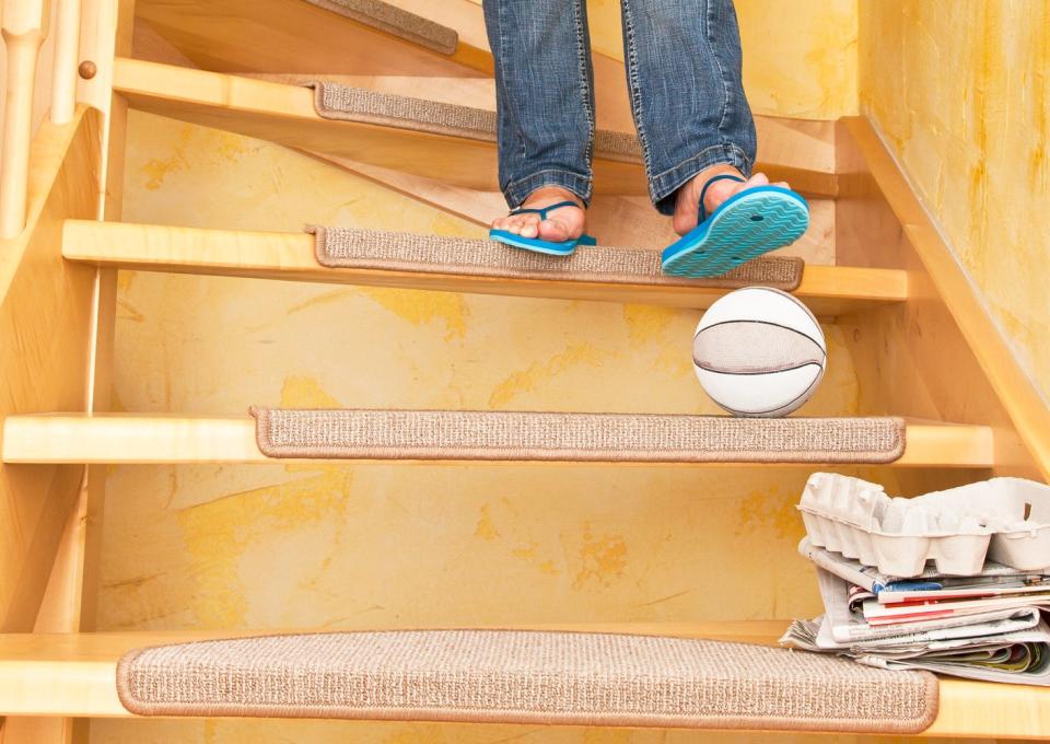 Person wearing jeans and flip-flops carrying a laundry basket up a staircase, with a soccer ball and newspapers on the steps. Faces not visible