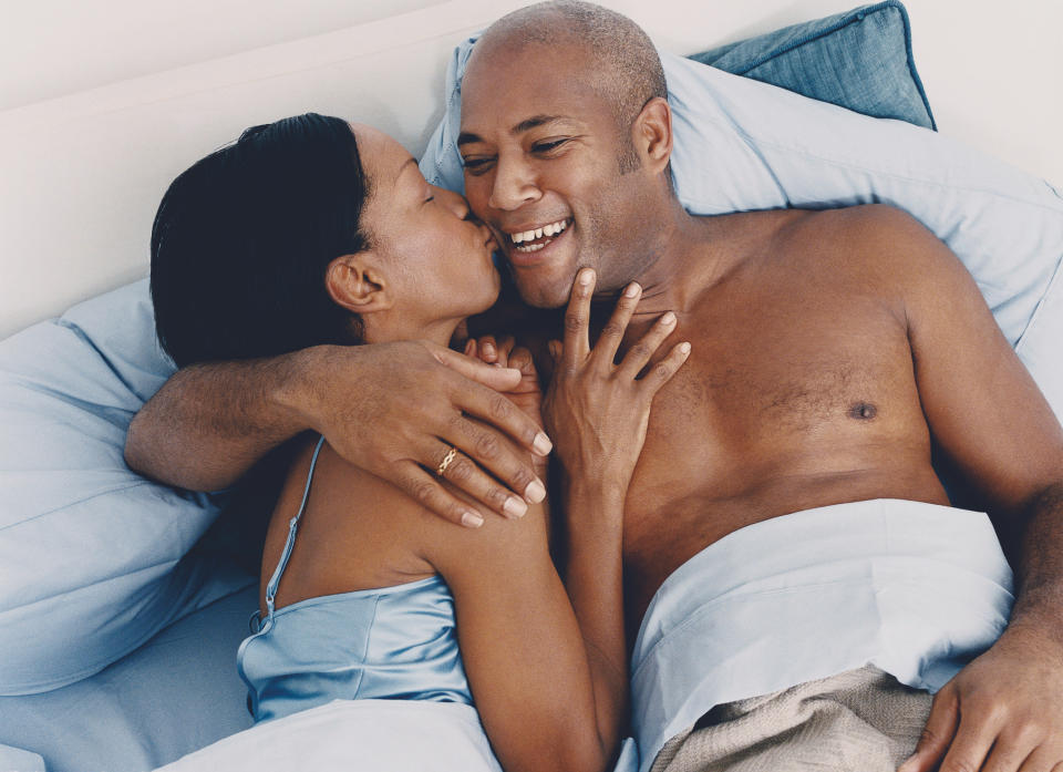 A woman kisses a shirtless man's cheek as they lie in bed, smiling and embracing each other. Both appear happy and relaxed. Names of persons unknown