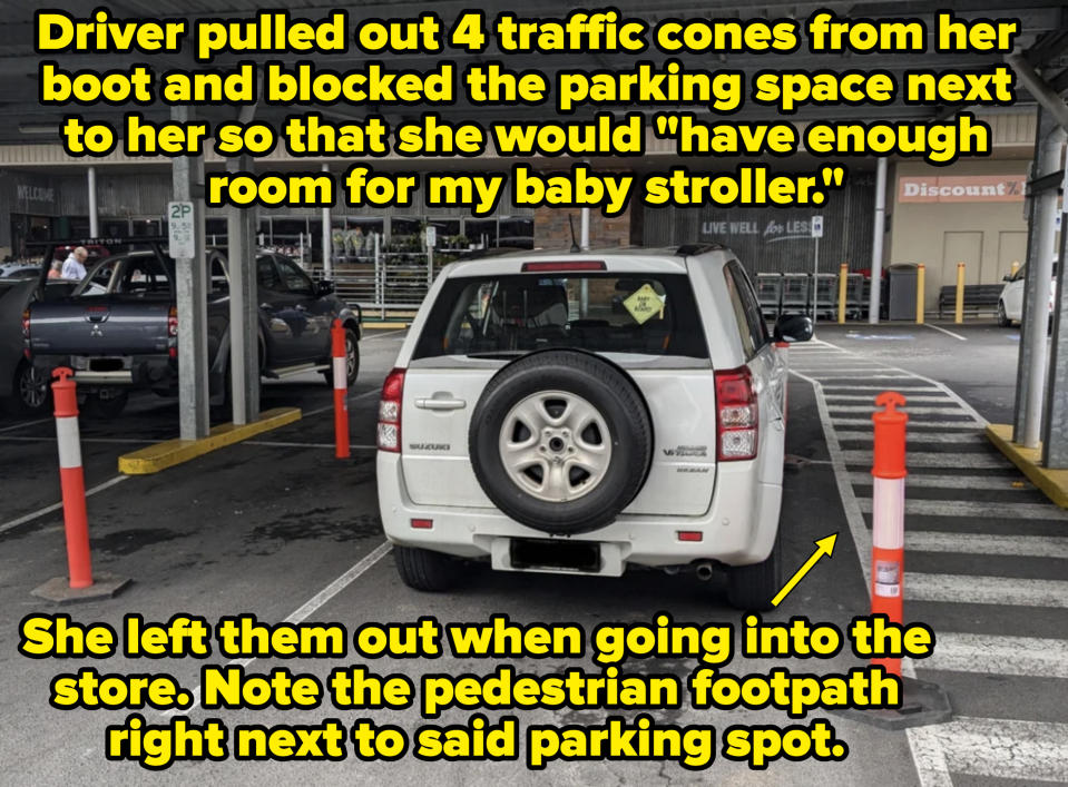 A white Suzuki SUV is parked in a covered parking spot with orange pylons around it in a partially filled lot outside a retail store