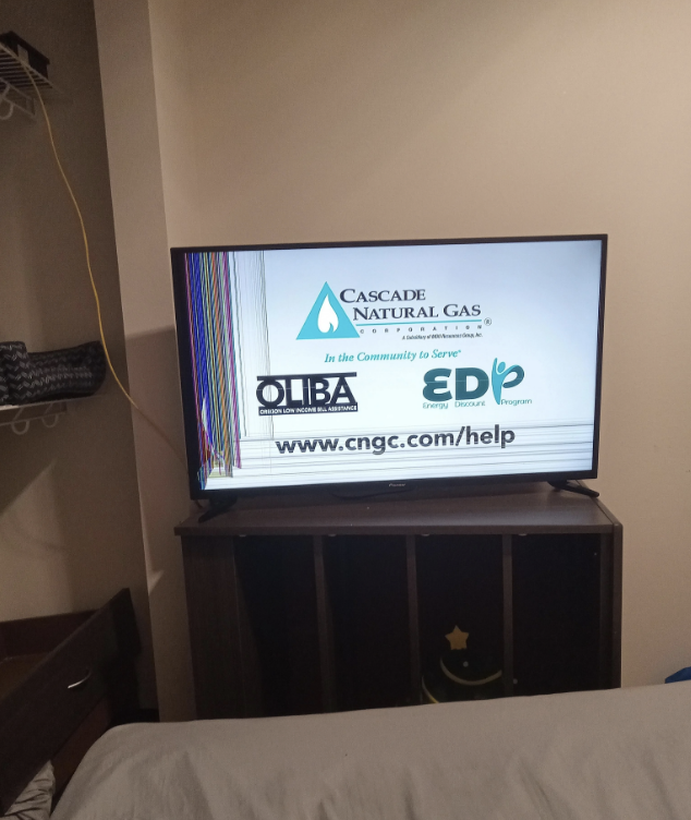 A TV displaying a Cascade Natural Gas ad with a damaged screen. The ad lists OIBA and Energy Direct Program logos and a help URL: www.cngc.com/help