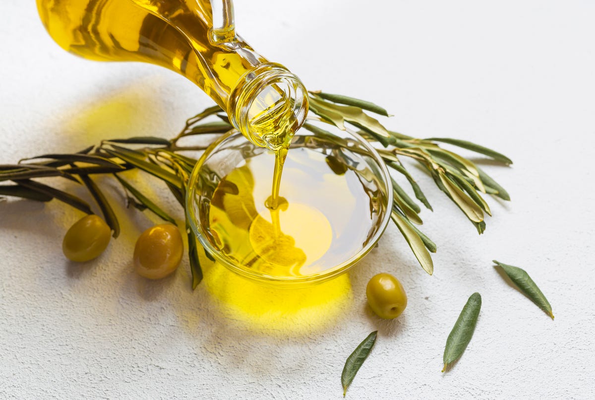 A glass jar of olive oil pouring into a glass bowl with an olive branch near.