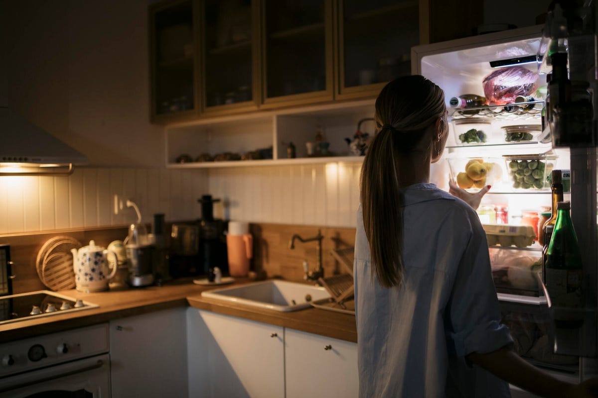 A woman in a dark kitchen opens a refrigerator filled with food.