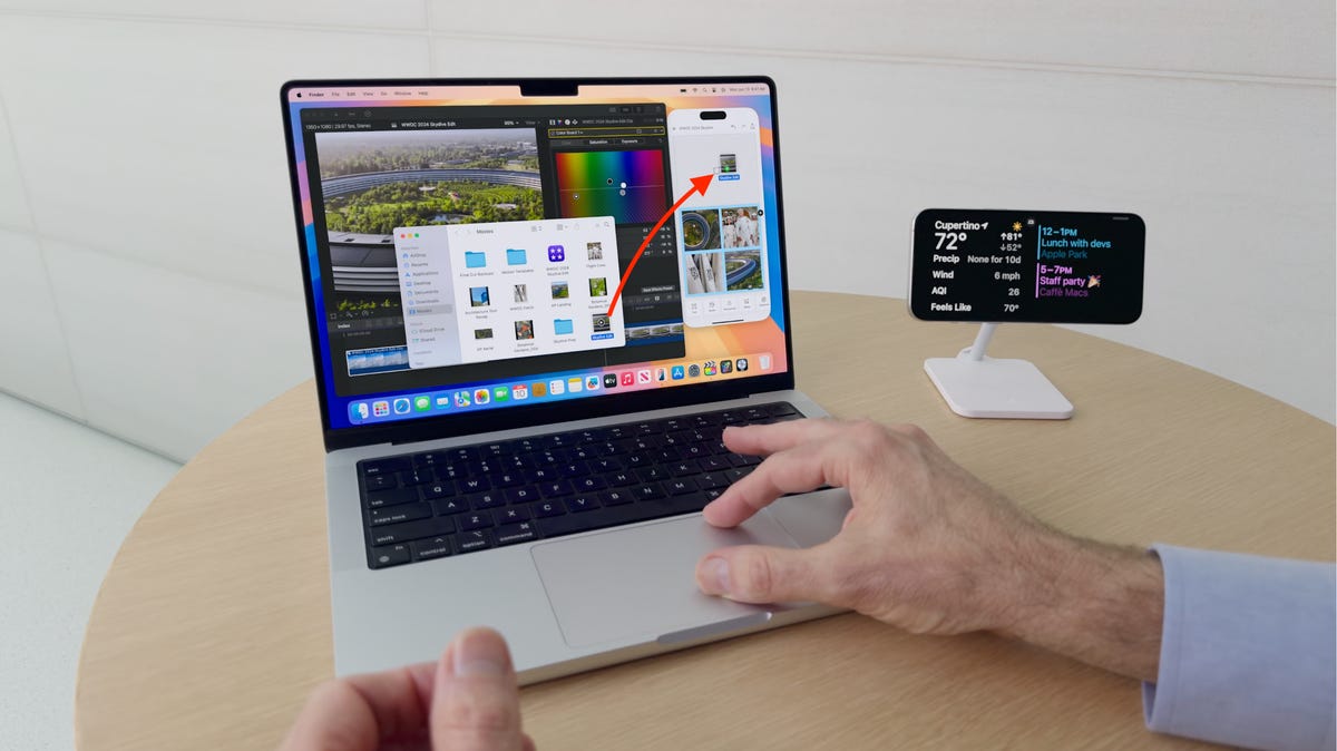A Mac laptop next to an iPhone on a stand, with hands using the trackpad to drag a file from the MacOS Finder to the iPhone via iPhone Mirroring.