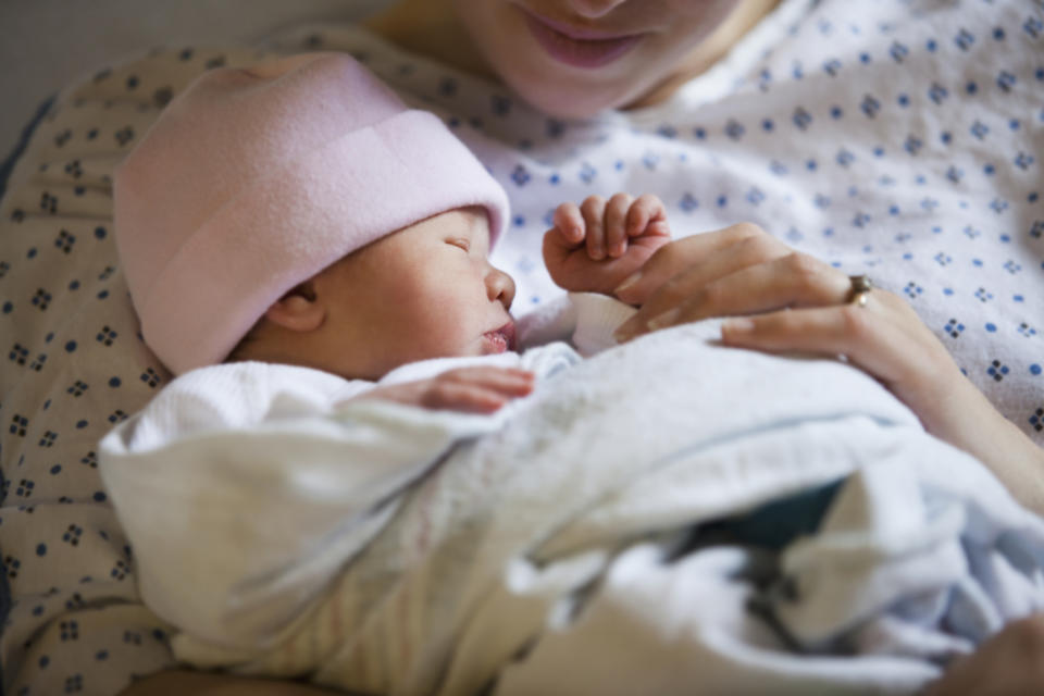 A newborn baby with a pink hat sleeps cradled in a woman's arms, who is wearing a hospital gown