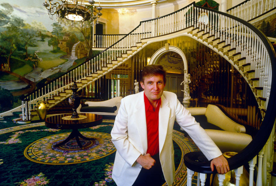 A young Donald Trump in a white blazer and red shirt stands in front of a grand staircase inside a luxurious mansion with elaborate decor