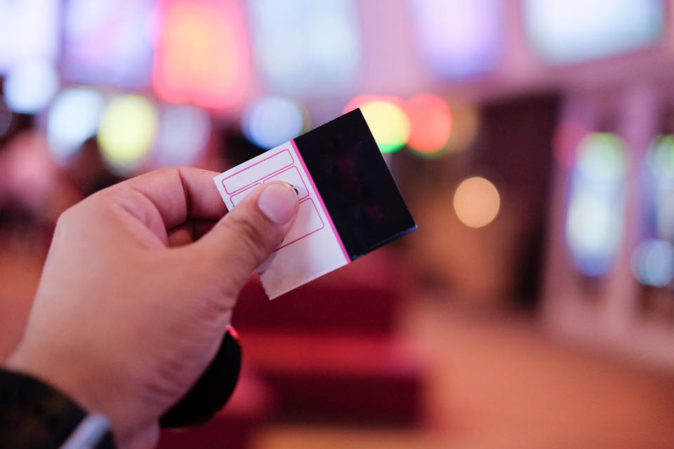 A hand holding a ticket in a brightly lit environment with blurred multicolored lights and screens in the background