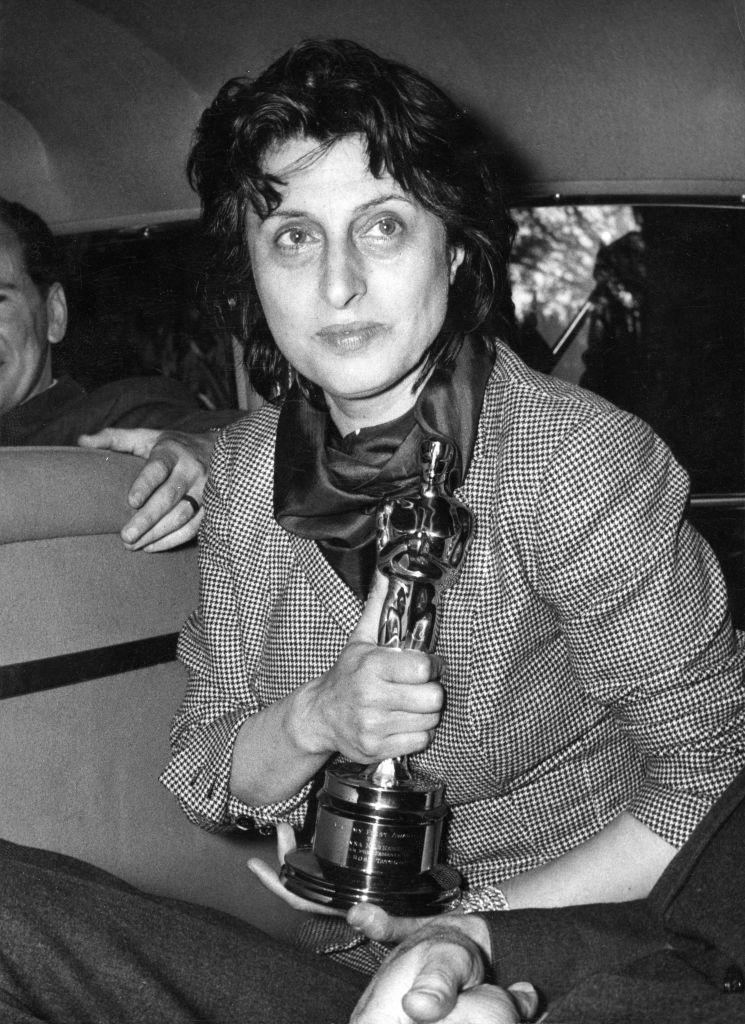 Anna Magnani holds an Oscar statuette while seated, wearing a checked suit jacket and a scarf around her neck