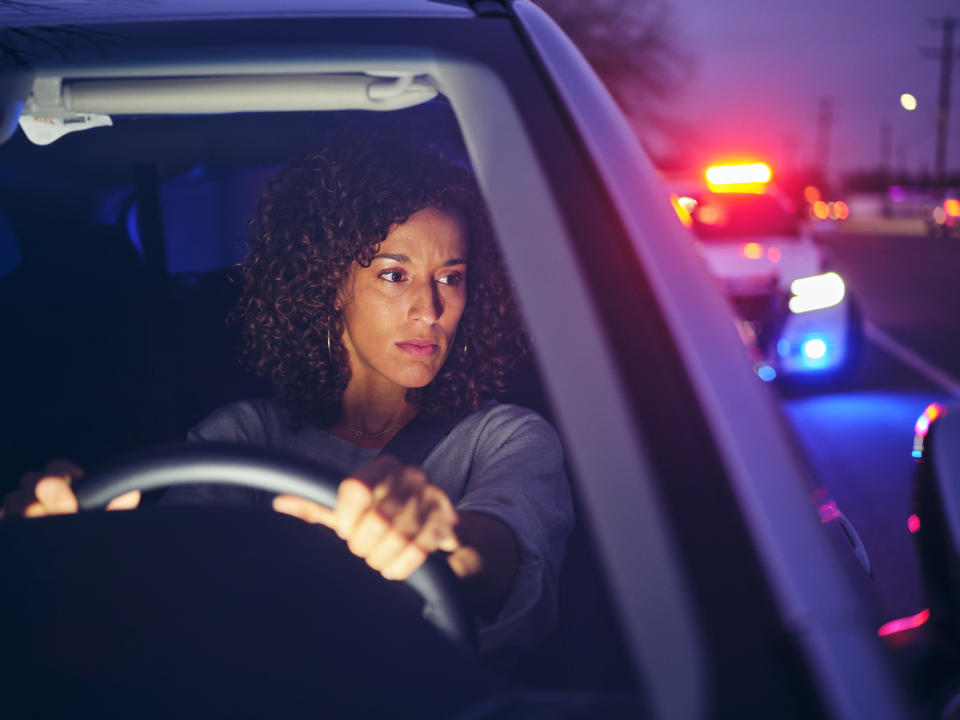 A woman with curly hair, driving a car at dusk, looks concerned as police car lights flash behind her