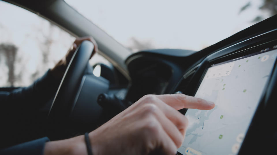 A hand is pointing at a GPS navigation screen in a car. The driver is holding the steering wheel