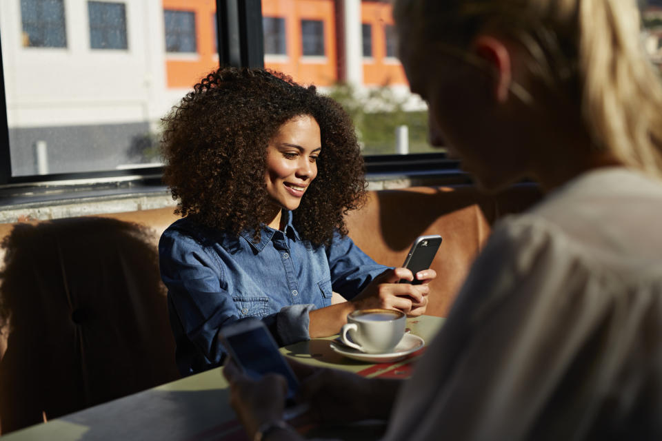Two people at a coffee shop, one smiling while using a phone, and another blurred in the foreground