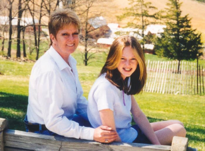 Two people sit outdoors on a wooden fence. The person on the left is older, with short hair, and wearing a white shirt. The person on the right is younger, with long hair