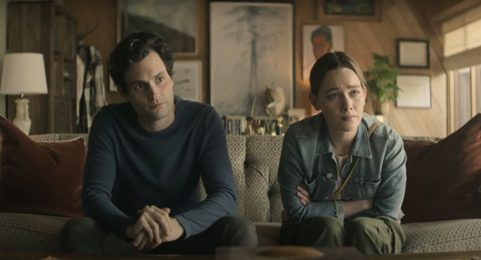 Penn Badgley and Victoria Pedretti sit on a couch, looking serious, with a cozy living room in the background