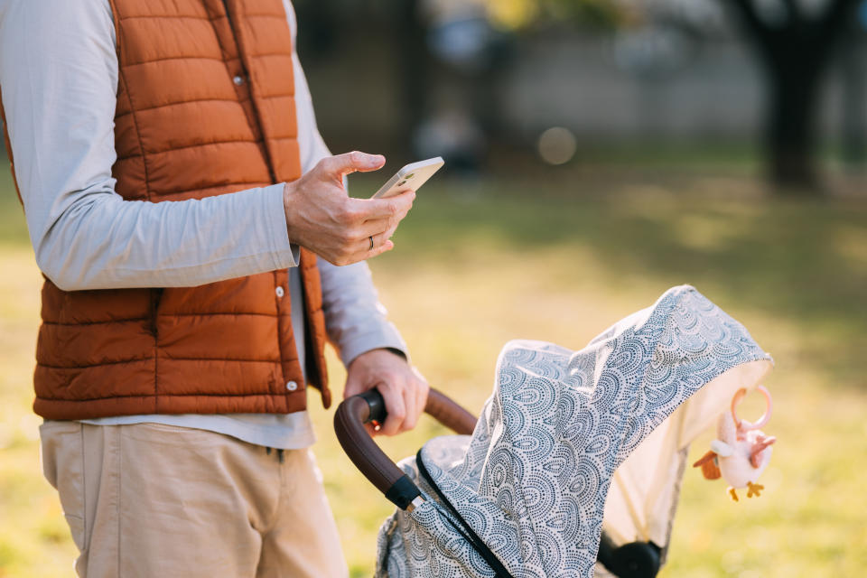 Person pushing a stroller with one hand, using a smartphone with the other hand, outdoors on a sunny day. The stroller has a toy hanging from it