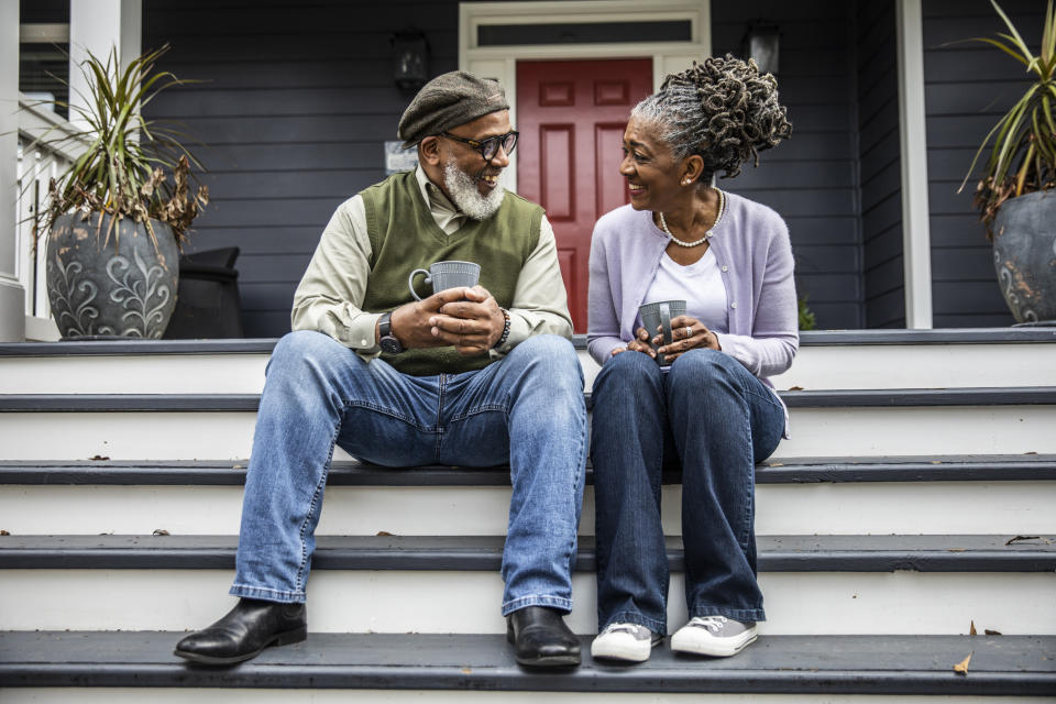 Older couple sitting on porch steps, both smiling and holding mugs. The man wears a hat, glasses, and casual clothing; the woman has gray dreadlocks and casual clothing