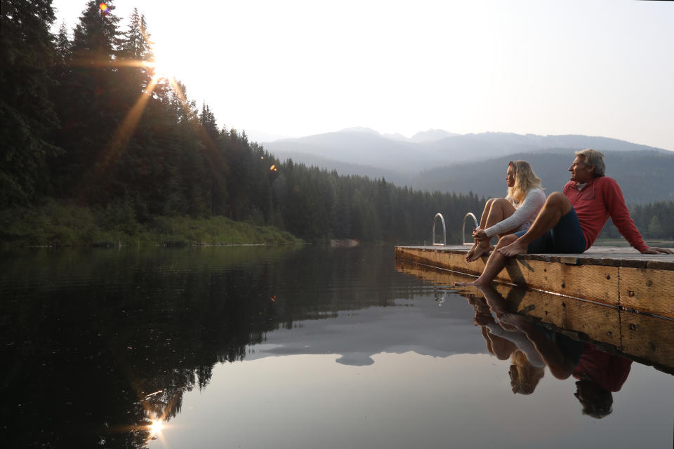 A woman and man sit at the edge of a wooden dock overlooking a serene lake surrounded by mountains and trees during sunrise or sunset