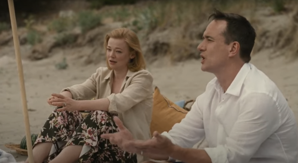 Sarah Snook and Matthew Macfadyen sit on a beach, talking under a beach umbrella while dressed in casual summer clothes
