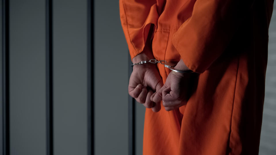 A person in an orange jumpsuit with hands cuffed behind their back, standing in front of prison bars