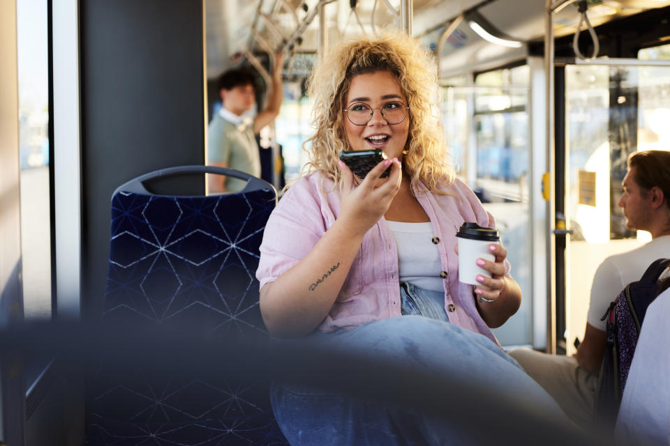 A woman with curly hair and glasses, wearing a casual shirt and overalls, is seated inside a bus. She is holding a coffee cup and speaking into her phone. Passengers are visible in the background