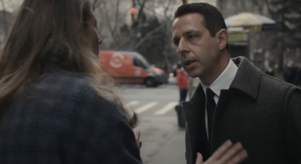 Jeremy Strong is in a heated conversation with another person on a city street. He is dressed in a formal coat and tie