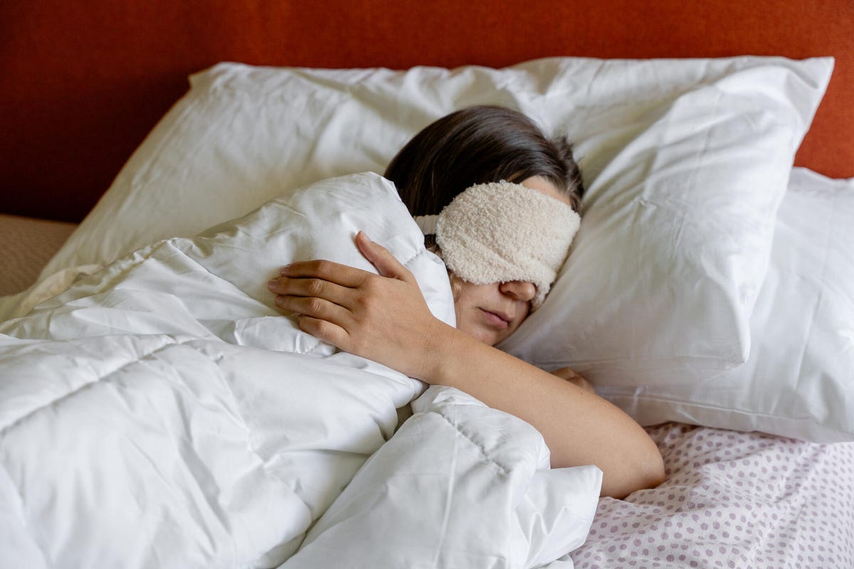A person sleeping in a bed with white bedding and a beige eye mask in front of a reddish wall.