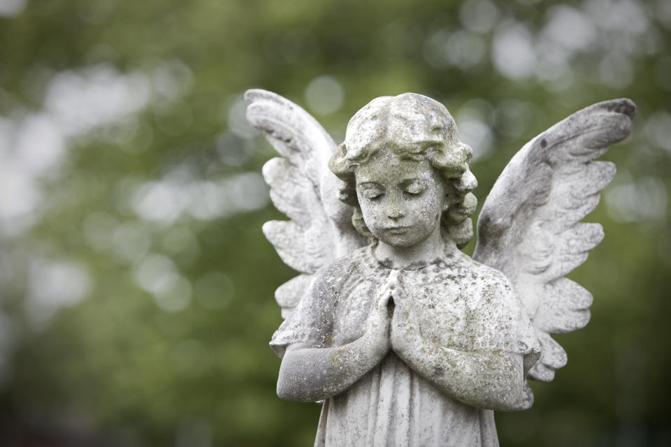 A stone angel statue with folded hands and closed eyes stands outdoors, surrounded by a blurred green background