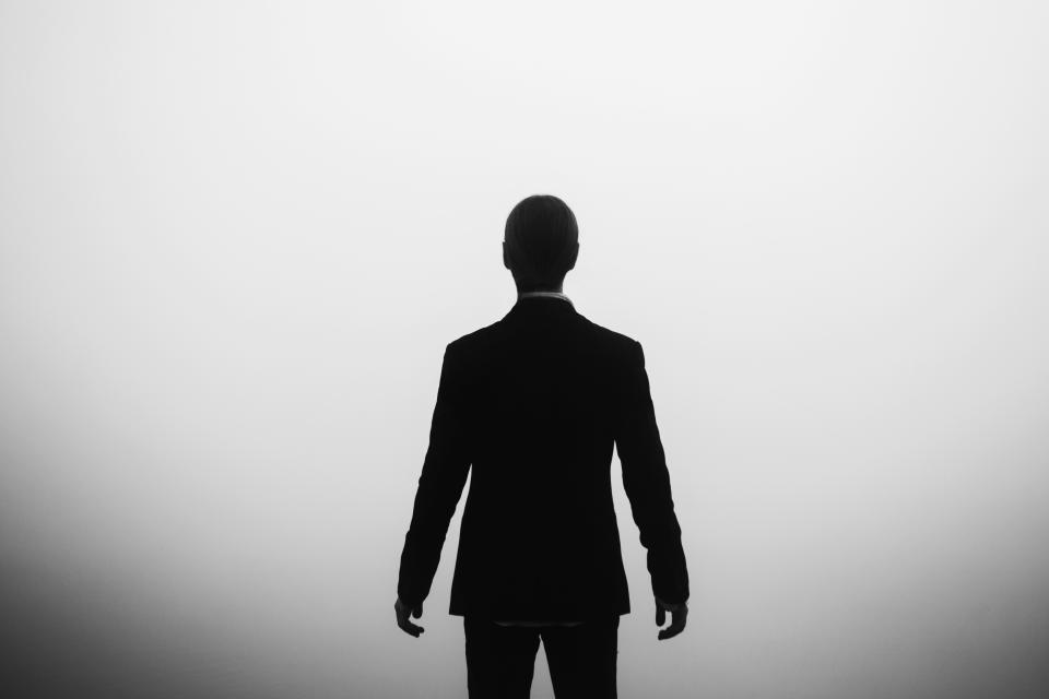 Silhouette of a person in a suit standing facing an empty, faded background. No distinguishable features or specific details are visible