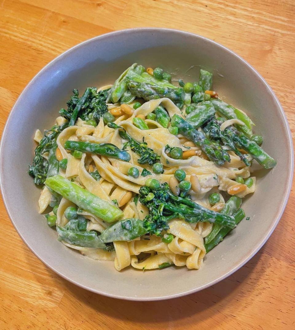Bowl of creamy pasta with asparagus, peas, pine nuts, and leafy greens on a wooden table