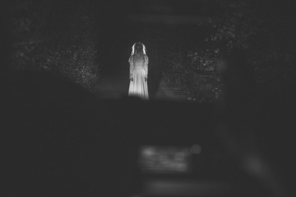 A mysterious figure in a long dress is illuminated at night, standing on a dark road. The figure appears ghostly and ethereal, adding an eerie atmosphere
