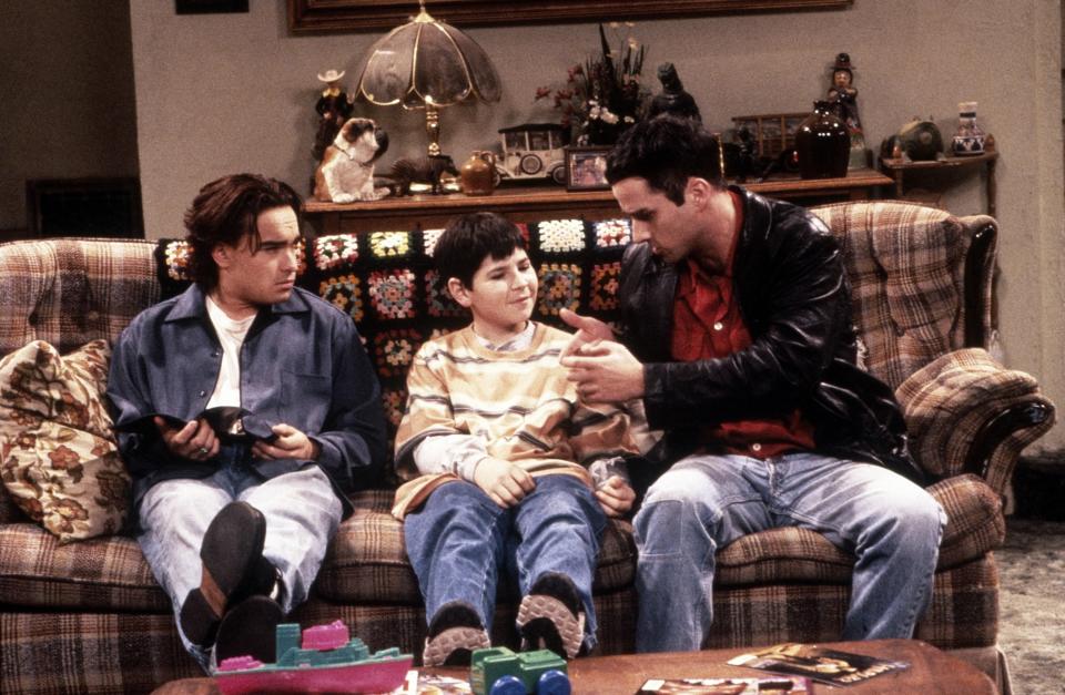 Three people sit on a couch; two adults with casual 90s style, and a child in a striped sweater between them, in a cozy, vintage living room setting
