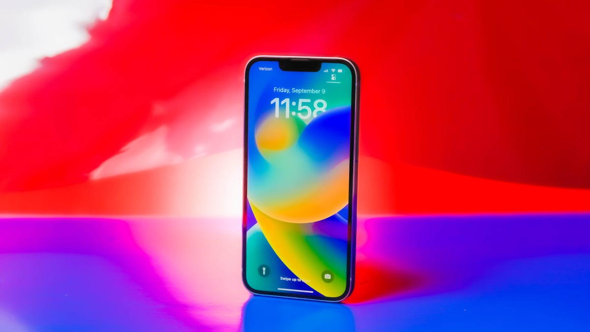 The iPhone 14 sits on a red and blue background, with a blue, orange, green and yellow wallpaper lit up on the screen of the phone.