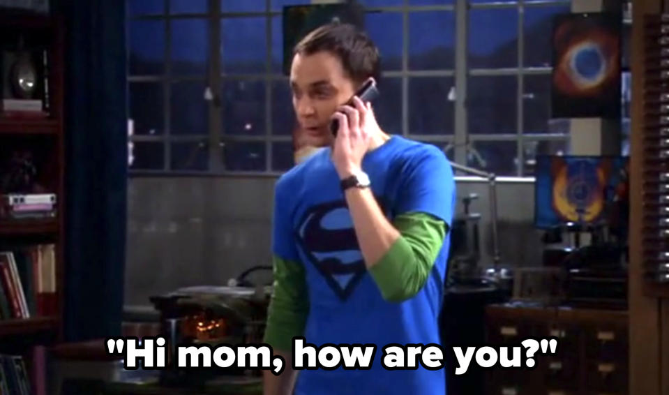 A person wearing a blue Superman t-shirt with a green long-sleeve shirt underneath is talking on the phone in a room with large windows