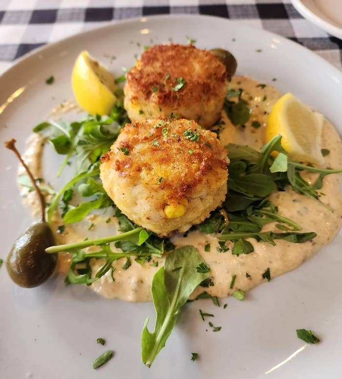 Two crab cakes on arugula with sauce, garnished with lemon wedges and capers, on a plate
