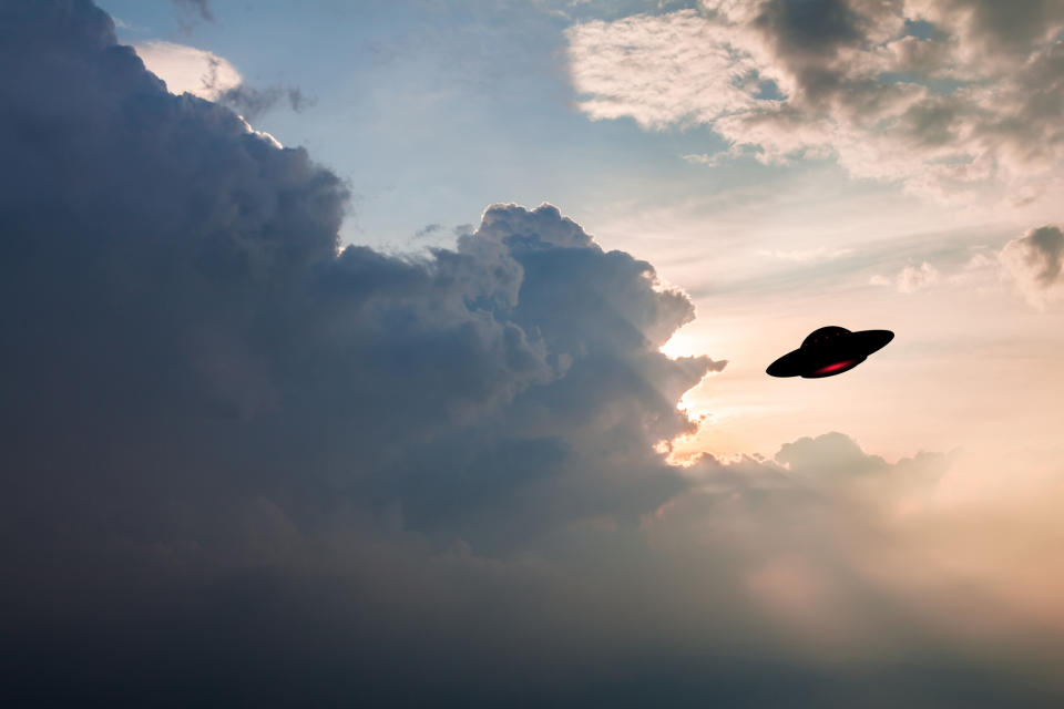 Silhouette of a UFO flying against a dramatic sky with clouds and sunlight peeking through