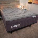 The Purple mattress on a black bed frame in a warmly lit room.