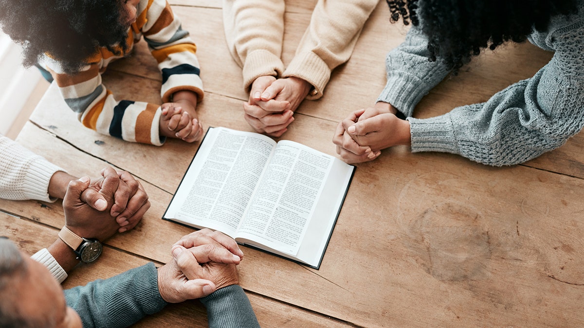 A multigenerational group prays around an open Bible at a wooden table
