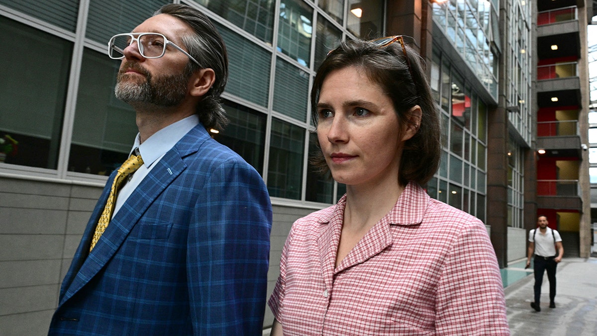 Amanda Knox wearing a pink patterned shirt walking alongside a man in a blue plaid suit.