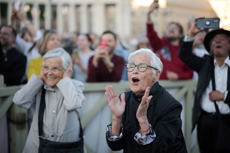 Two elder women clap and hoot — one shielding her ears from the noise — as a new pope is announced at the Vatican